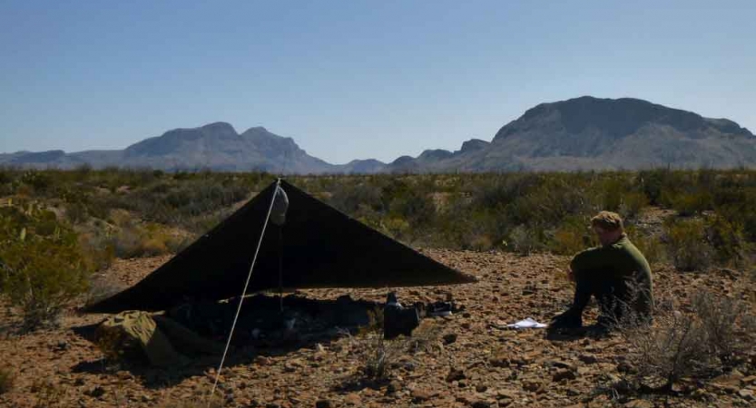 a student sits beside their tarp shelter in the desert with mountains in the background